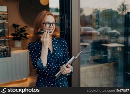 Smiling pensive redhead young woman makes voice call via smartphone looks away stands in door of coffee shop uses modern technologies for communication holds laptop and diary dressed elegantly. Young woman makes voice call uses modern technologies for communication