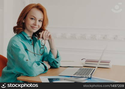 Smiling pleased young red head woman ready for brainstroming with colleagues online, poses in coworking space with paper documents, going to participate in online conference, works from home