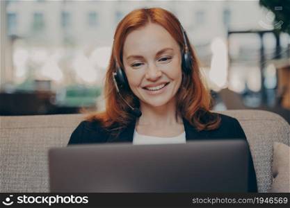 Smiling pleased young redhead woman wearing headset talking with colleagues online on laptop while sitting on sofa in modern office, participating in online conference during remote work from home. Smiling pleased young redhead woman wearing headset talking with colleagues online on laptop