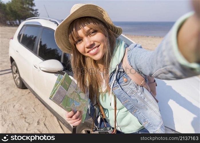 smiling portrait female traveler holding map hand taking selfie with her car beach