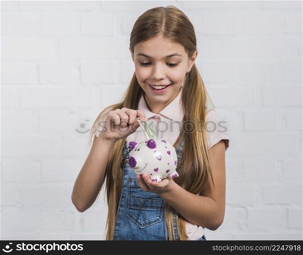 smiling portrait girl inserting currency note white piggybank