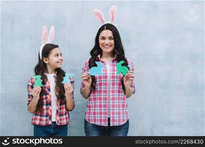 smiling portrait mother daughter holding paper cutout bunny against blue wall