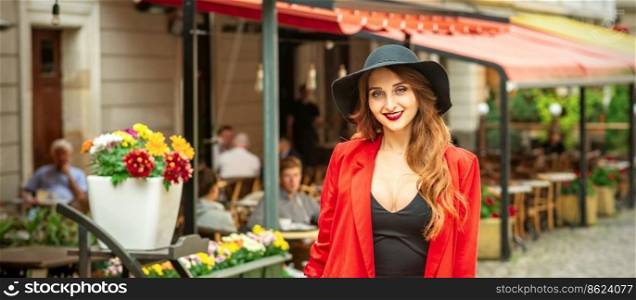 Smiling portrait of fashion young caucasian woman is posing in black hat and red jacket in the city street. Woman is posing in the city street