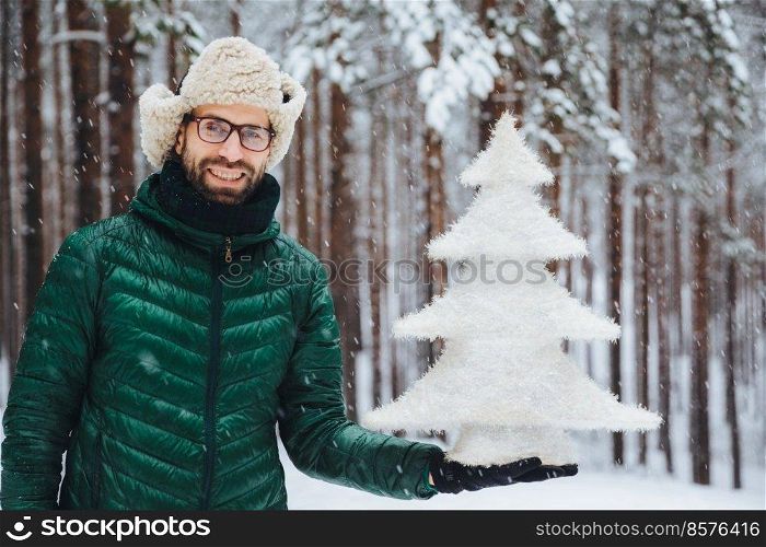 Smiling positive male wears glasses, hat and anorak, holds artificial fir tree, stands against trees covered with snow, looks directly into camera with happy expression, spends time on frosty weather