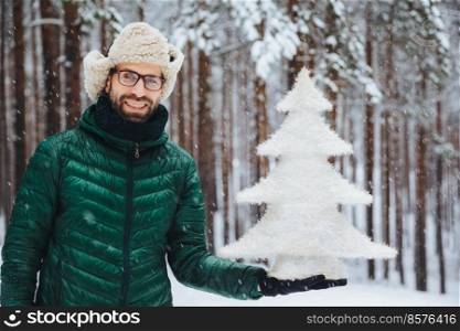 Smiling positive male wears glasses, hat and anorak, holds artificial fir tree, stands against trees covered with snow, looks directly into camera with happy expression, spends time on frosty weather