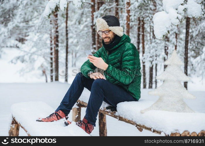 Smiling positive male wears warm winter clothes, reads message on mobile phone, spends free time in calm atmosphere outdoors in winter forest, enjoys fresh air. People, rest, technology concept