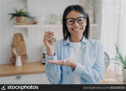 Smiling positive spanish woman is showing house key in her hand. Success and achievement concept. Happy real estate owner. Mortgage loan and investment. Modern scandinavian interior of kitchen.. Smiling positive spanish woman is showing house key in her hand. Happy real estate owner.