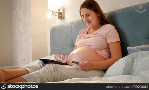 Smiling pregnant woman doing online shopping on tablet computer before going to sleep at night. Smiling pregnant woman doing online shopping on tablet computer before going to sleep at night.