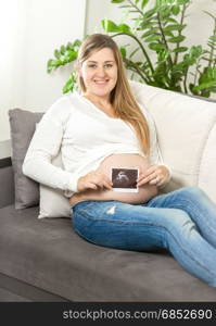 Smiling pregnant woman sitting on sofa and holding ultrasound fetus scan