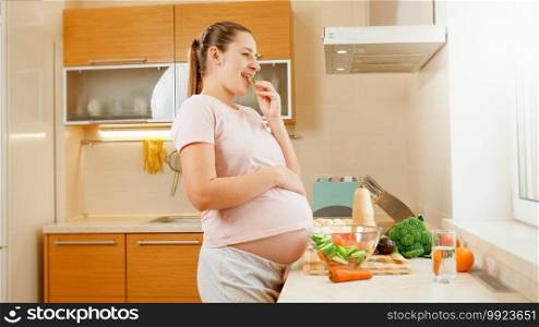 SMiling pregnant woman waiting for baby eating fresh vegetables and looking out of window on kitchen. Concept of healthy lifestyle and nutrition during pregnancy.. SMiling pregnant woman waiting for baby eating fresh vegetables and looking out of window on kitchen. Concept of healthy lifestyle and nutrition during pregnancy