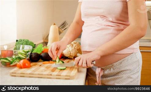SMiling pregnant wooman making salad and eating fresh cucumber on kitchen at house. Concept of healthy lifestyle and nutrition during pregnancy.. SMiling pregnant wooman making salad and eating fresh cucumber on kitchen at house. Concept of healthy lifestyle and nutrition during pregnancy