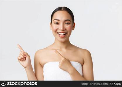 Smiling pretty asian girl in towel pointing fingers upper left corner as advertise spa salon, beauty product, recommend cleanser or skincare cosmetics, standing white background.. Smiling pretty asian girl in towel pointing fingers upper left corner as advertise spa salon, beauty product, recommend cleanser or skincare cosmetics, standing white background
