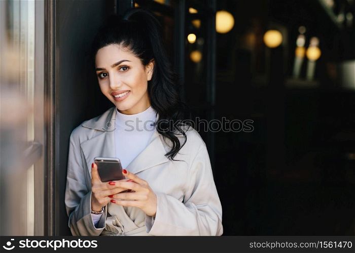 Smiling pretty brunette woman with pony tail having red manicure dressed formally holding smartphone doing online shopping while waiting for train. Businesswoman with cell phone going for work
