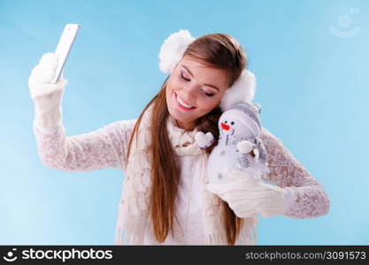 Smiling pretty cute woman holding little snowman taking selfie self photo picture with camera. Attractive girl in earmuffs and white sweater pullover in studio on blue. Winter fashion.. Woman with little snowman taking selfie photo.