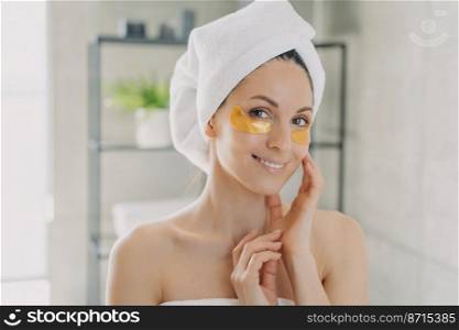 Smiling pretty hispanic woman wearing towel using golden moisturizing hydrogel patches on under eye skin in bathroom. Female enjoying morning antiaging skincare routine after shower.. Smiling woman wearing towel using hydrogel patches on under eye skin in bathroom. Skincare routine