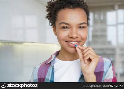 Smiling pretty mixed race young girl holding a pill, looking at camera indoors, taking daily medicine, vitamin D, dietary supplements for healthy skin, hair, nails and beauty. Head shot portrait.. Smiling mixed race young girl holding pill takes medicine, vitamin, supplements for beauty, headshot