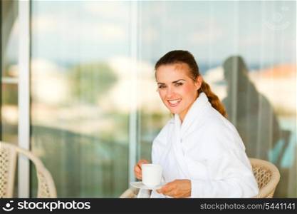 Smiling pretty woman in bathrobe sitting at table on terrace and having cup of tea&#xA;