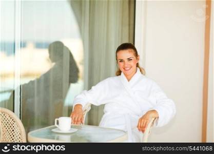 Smiling pretty woman in bathrobe sitting at table on terrace &#xA;