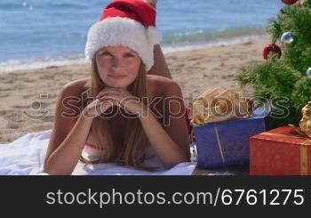 Smiling pretty woman in Santa hat with gifts under decorated Christmas tree on tropical beach