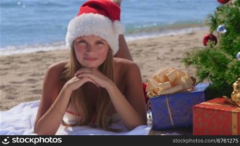 Smiling pretty woman in Santa hat with gifts under decorated Christmas tree on tropical beach