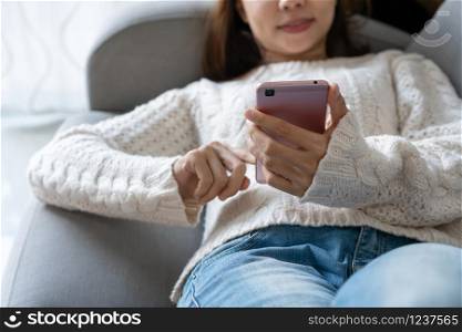 Smiling pretty woman in white sweater using mobile phone while lying down on a sofa in the living room at home