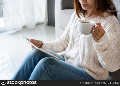 Smiling pretty woman in white sweater using tablet computer while holding a cup of coffee sitting in the living room at home