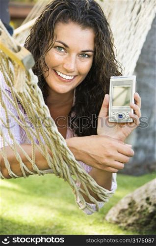 Smiling pretty young adult Caucasian brunette female lying in hammock holding up PDA towards viewer.