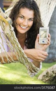 Smiling pretty young adult Caucasian brunette female lying in hammock holding up PDA towards viewer.