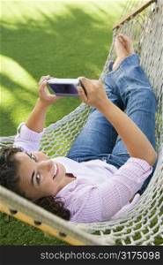 Smiling pretty young adult Caucasian brunette female lying in hammock looking up at veiwer and using PDA.