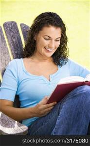 Smiling pretty young adult Caucasian brunette female sitting in chair reading book.