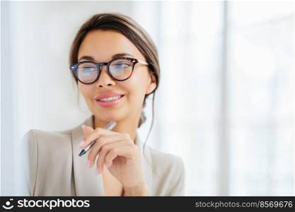 Smiling prosperous lady work in business sphere, holds pen, wears elegant clothes, has healthy skin, minimal makeup, looks somewhere, poses indoor against blurred white background, copy space left