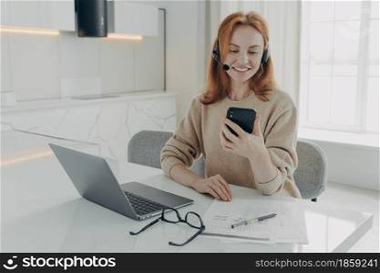 Smiling red-haired european female office worker in headset using smartphone while sitting at her workplace with laptop, happy woman talking by video call on mobile phone during remote work at home. Smiling female office worker in headset using smartphone while sitting at her workplace with laptop