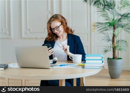 Smiling red haired female focused in modern cellular, happy to recieve text message, poses at workplace, drinks coffee, works on financial project gets ready for video conference with business partner