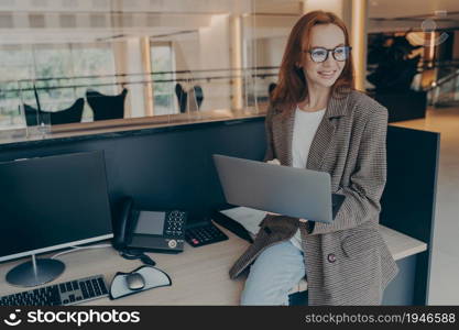 Smiling red-haired female office employee in glasses sitting on desk with various equipment while holding laptop, looking around and surfing web on computer while working in modern coworking space. Smiling female office employee sitting on desk at her workplace while holding laptop