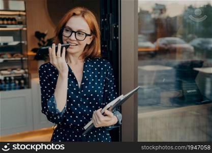 Smiling redhead female freelancer with laptop stands outdoors with mobile phone and uses voice recognition system on smartphone, leaving coffee shop and records audio message on modern cellphone. Female freelancer stands outdoors with mobile phone and uses voice recognition system on smartphone