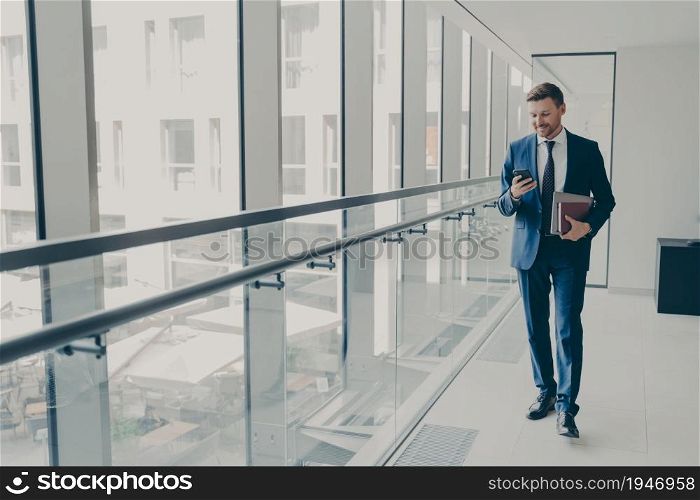 Smiling redhead man office worker in suit using cellphone, checking email or news feed of social network account with laptop and notebook in hand, looking at smartphone while standing in office. Smiling redhead man office worker in suit using cellphone while standing in modern office