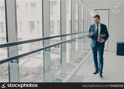 Smiling redhead man office worker in suit using cellphone, checking email or news feed of social network account with laptop and notebook in hand, looking at smartphone while standing in office. Smiling redhead man office worker in suit using cellphone while standing in modern office