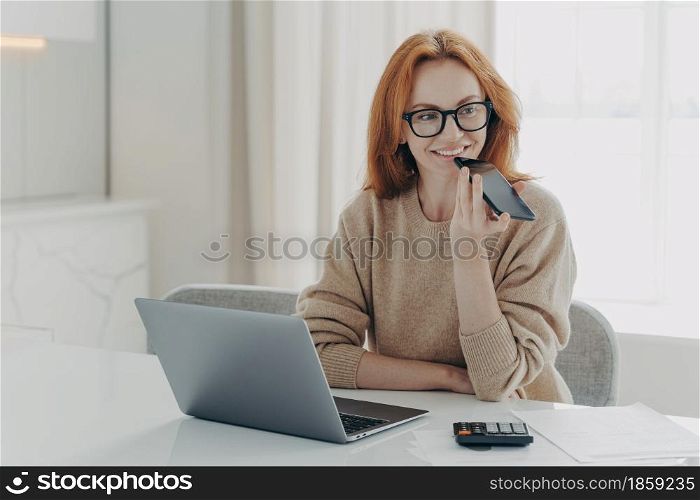 Smiling redhead woman accountant financier uses voice recognition system on smartphone, records audio message on modern cellphone while working remotely on laptop at home or office, wearing spectacles. Smiling woman accountant financier uses voice recognition system on smartphone, records audio message