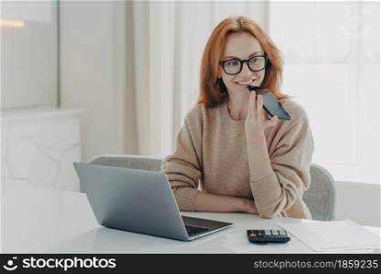 Smiling redhead woman accountant financier uses voice recognition system on smartphone, records audio message on modern cellphone while working remotely on laptop at home or office, wearing spectacles. Smiling woman accountant financier uses voice recognition system on smartphone, records audio message