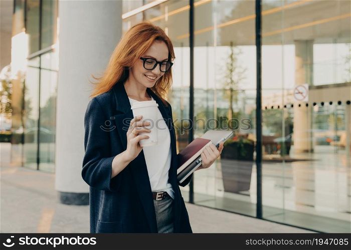 Smiling redhead woman corporate worker dressed in formal clothes drinks coffee carries notebook for making notes laptop focused down with happy expression poses near entrance of office building. Smiling redhead woman corporate worker dressed in formal clothes drinks coffee