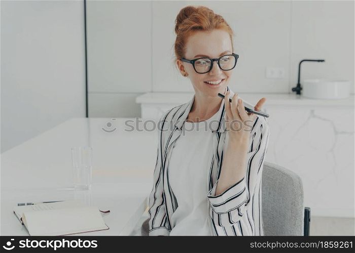 Smiling redhead woman uses virtual voice assistant turns away focused into distance records audio message wears spectacles striped shirt sits at white desk makes notes in notepad works at home. Smiling redhead woman uses virtual voice assistant records audio message