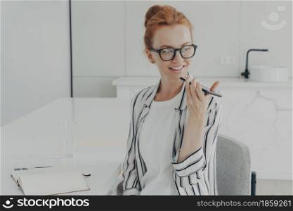 Smiling redhead woman uses virtual voice assistant turns away focused into distance records audio message wears spectacles striped shirt sits at white desk makes notes in notepad works at home. Smiling redhead woman uses virtual voice assistant records audio message