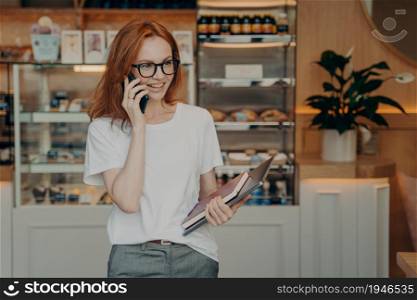 Smiling redhead young woman has telephone conversation enjoys positive talk carries notebook and laptop computer poses against bakery shop background. People communication lifestyle concept.. Smiling redhead young woman has telephone conversation enjoys positive talk