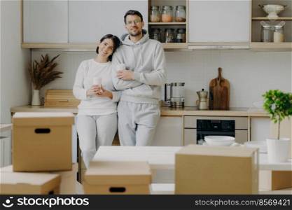 Smiling relaxed husband and wife drink coffee at kitchen, hold paper cups with hot beverage, dressed in casual outfit, relocate in new apartment, pile of cardboard boxes with property in foreground.