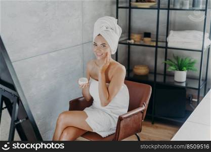 Smiling relaxed woman cares of her beautiful skin, sits in comfortable armchair, poses in cozy bathroom, looks at herself in mirror, holds jar of skin cream, touches cheek gently, wrapped in towel