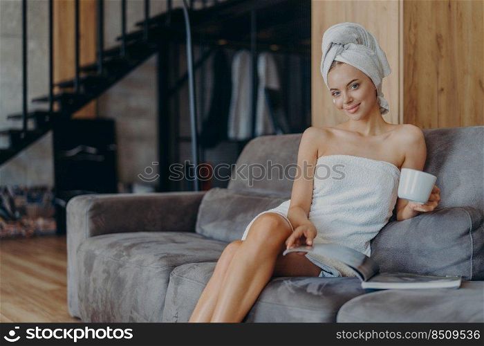 Smiling relaxed young European woman sits on comfortable sofa, reads magazine and drinks coffee, poses on comfortable sofa, undergoes beauty treatments, has satisfied expression. Spending time at home