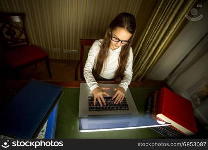 Smiling schoolgirl in eyeglasses working at computer at dark room