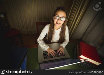 Smiling schoolgirl in white shirt and eyeglasses working at computer at dark room