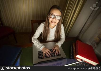 Smiling schoolgirl in white shirt doing homework at laptop in dark room