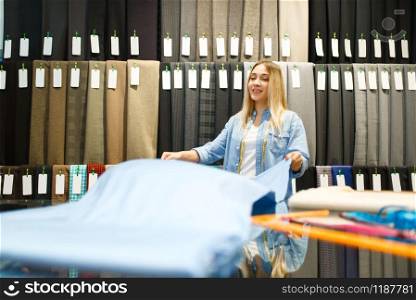 Smiling seamstress holds fabric in textile store. Shelf with cloth for sewing on background, clothing patterns choice in shop. Smiling seamstress holds fabric in textile store